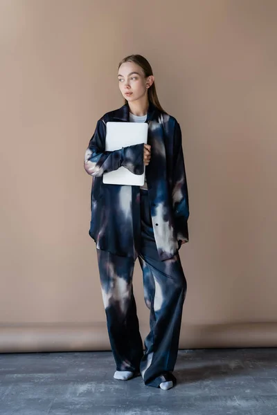 Повна довжина жінки в модному градієнтному одязі, що стоїть з ноутбуком на бежевому фоні — стокове фото
