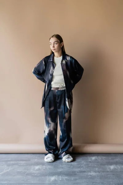 Full length of woman in oversize clothes and soft slippers posing with hands behind back on beige background — Foto stock