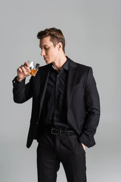 Mann im schwarzen Anzug steht mit der Hand in der Tasche und trinkt Whisky isoliert auf grau — Stockfoto
