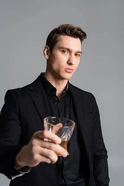 Confident man in formal wear toasting with blurred glass of whiskey isolated on grey - foto de stock