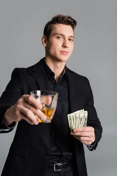 Confident man holding dollar banknotes and toasting with blurred glass of whiskey isolated on grey - foto de stock