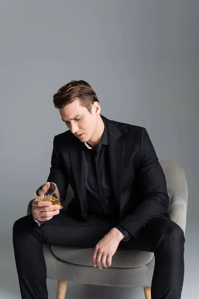 Thoughtful man with glass of whiskey sitting in armchair isolated on grey — Stock Photo