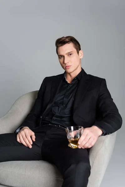 Serious man in black suit sitting in armchair with glass of whiskey isolated on grey — Stockfoto