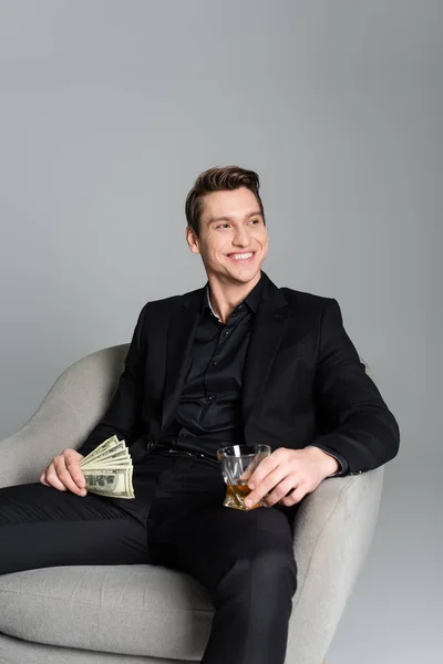 Счастливый человек с деньгами и виски, сидящий в кресле и уединяющийся на сером — стоковое фото