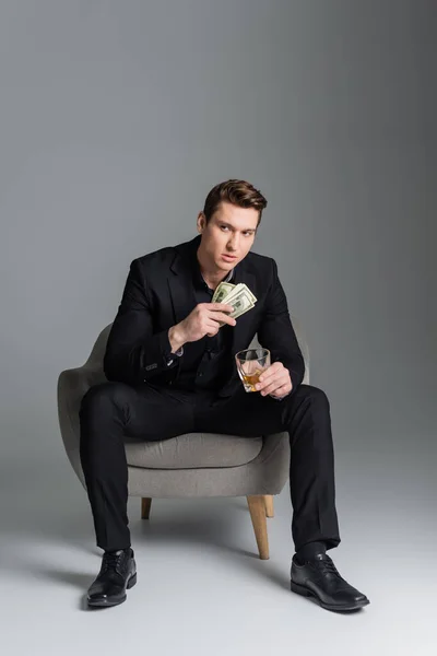 Full length of man in black suit sitting in armchair with money and whiskey on grey background — Fotografia de Stock