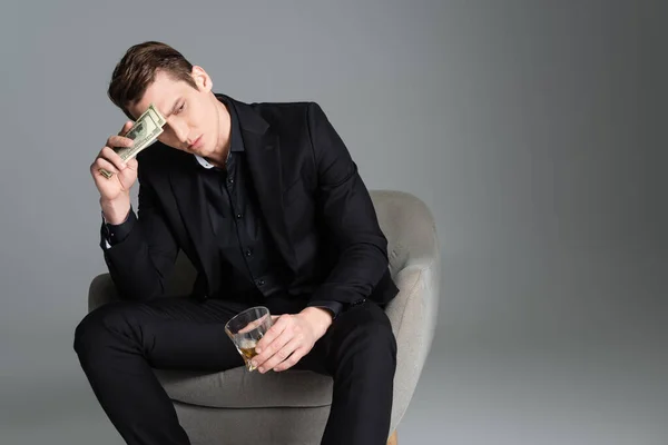 Thoughtful man with glass of whiskey and money sitting in armchair and looking away isolated on grey — Photo de stock