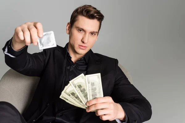 Man in black formal wear holding condom and dollar banknotes on blurred foreground isolated on grey - foto de stock