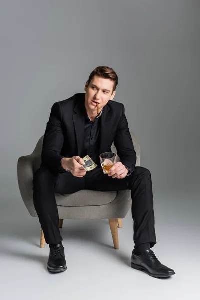 Full length of man with cigar and whiskey holding dollars and condom while sitting on grey background - foto de stock