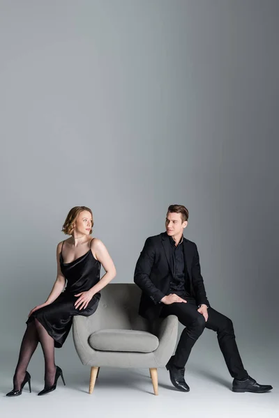 Volle Länge des Paares in schwarzen Kleidern, die einander auf einem Sessel auf grauem Hintergrund anschauen — Stockfoto