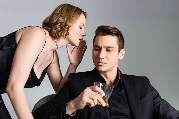 Сексуальная женщина шепчет на ухо мужчине со стаканом виски, изолированным на сером — стоковое фото