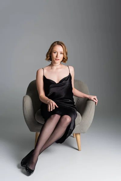 Повна довжина красивої жінки в чорній сукні ремінця, що сидить у кріслі на сірому фоні — стокове фото