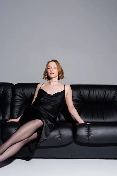 Mujer sexy en vestido de correa negro sentado en el sofá de cuero y mirando a la cámara aislada en gris - foto de stock