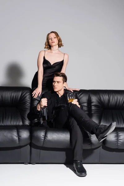 Sinnliche Frau blickt in die Kamera neben stilvollem Mann auf Couch sitzend mit Whisky auf grauem Hintergrund — Stockfoto