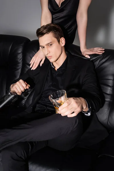 Mann mit Flasche und Glas Whiskey sitzt auf schwarzer Couch neben Frau auf grauem Hintergrund — Stockfoto