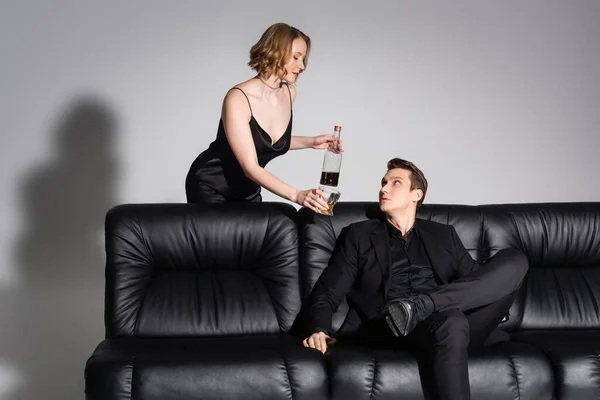 Sexy Frau mit Glas und Flasche Whiskey in der Nähe Mann sitzt auf schwarzer Couch auf grauem Hintergrund — Stockfoto