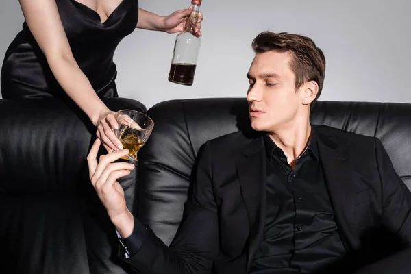 Woman proposing whiskey to man in black clothes sitting on leather sofa isolated on grey — Fotografia de Stock