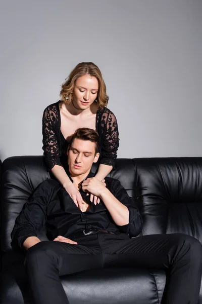 Verführerische Frau in Spitzenjacke verführt Mann sitzt auf schwarzem Sofa isoliert auf grau — Stockfoto
