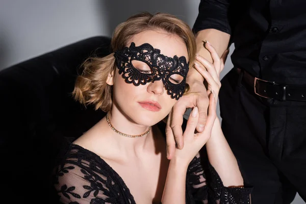 Sensual woman in black lace mask seducing man and holding his hand on grey background — Fotografia de Stock