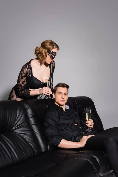 Соблазнительная женщина с бокалом шампанского рядом с мужчиной на черном кожаном диване, изолированном на сером — стоковое фото