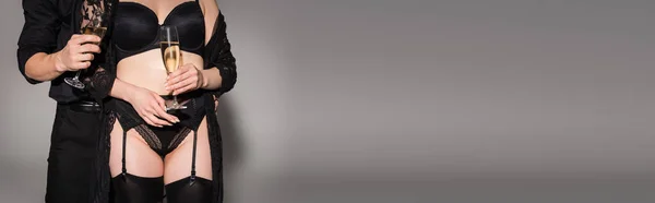 Vue partielle de femme sexy en lingerie et homme en noir tenant des verres à champagne isolés sur gris, bannière — Photo de stock