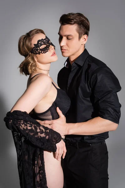 Uomo in camicia nera che abbraccia donna sexy in maschera di pizzo e reggiseno su sfondo grigio — Foto stock