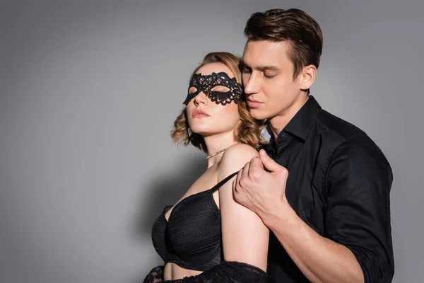 Man with closed eyes hugging sexy woman in black lace mask on grey background — Stock Photo