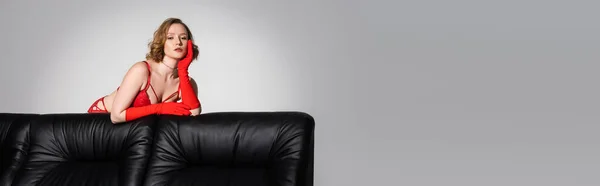 Sensuale giovane donna in reggiseno rosso e guanti guardando la fotocamera vicino al divano nero isolato su grigio, banner — Foto stock