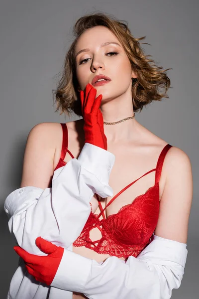Young and sexy woman in red bra and gloves touching chin and looking at camera isolated on grey - foto de stock