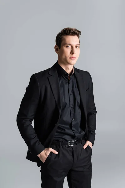 Confident man in black suit standing with hands in pockets and looking at camera isolated on grey - foto de stock