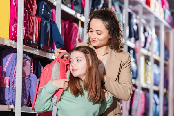 Sorrindo mulher tentando nova mochila na filha em papelaria — Fotografia de Stock