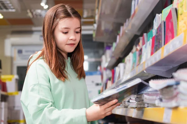 Schoolgirl looking at new copybook near blurred rack in stationery shop - foto de stock