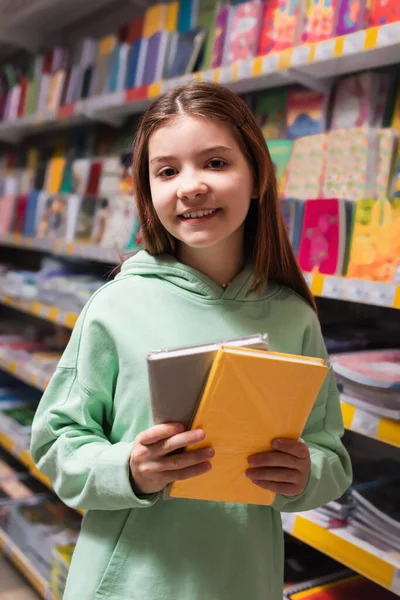Pleased girl with colorful notebooks smiling at camera in stationery store — Stock Photo