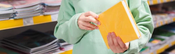 Partial view of child holding notebook near blurred rack in shop, banner - foto de stock