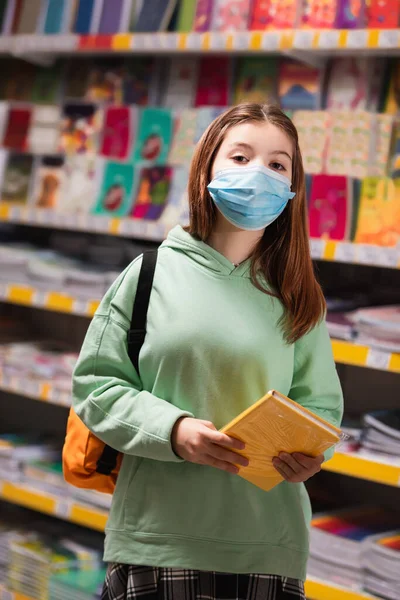 Girl in medical mask holding notebooks near blurred rack with school supplies — Stock Photo