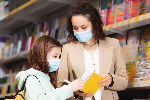 Woman in medical mask showing new notebook to daughter in stationery store — Stock Photo