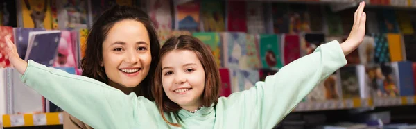 Cheerful woman and excited girl with outstretched hands looking at camera in stationery store, banner — Stock Photo