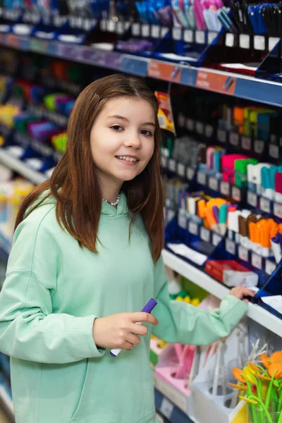 Усміхнена дівчина з фетровою ручкою біля розмитої стійки зі шкільними приладдям в канцелярському магазині — стокове фото
