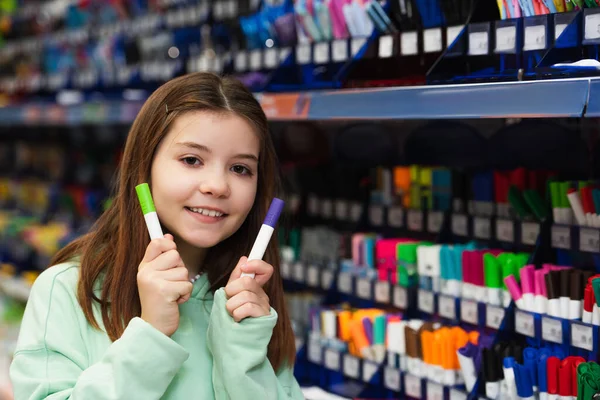 Усміхнена школярка тримає барвисті повстяні ручки і дивиться на камеру в канцелярському магазині — стокове фото