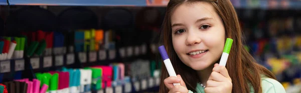 Happy schoolgirl with felt pens smiling at camera in stationery store, banner — Photo de stock