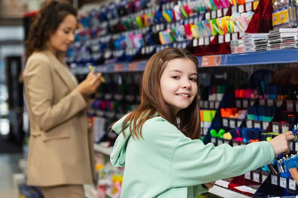 Happy schoolgirl smiling at camera near rack with school supplies and blurred mom — Photo de stock