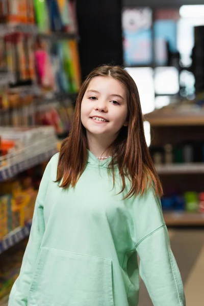 Щаслива школярка посміхається на камеру в розмитому канцелярському магазині — стокове фото