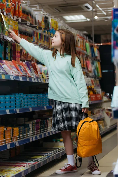Full length of schoolgirl with backpack choosing school supplies in stationery shop — стоковое фото
