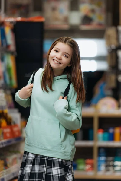 Pleased schoolgirl with backpack smiling at camera in stationery shop — Photo de stock