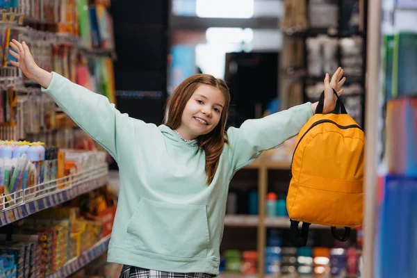 Весела дівчина з рюкзаком і розтягнутими руками дивиться на камеру в канцелярському магазині — стокове фото