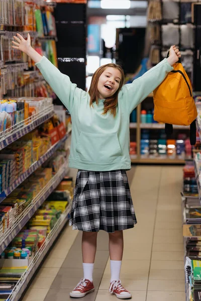 Happy girl in plaid skirt standing with outstretched hands and backpack in stationery store — Photo de stock
