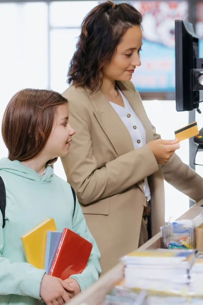 Woman holding credit card near payment terminal and daughter with copybooks in stationery store — Stockfoto