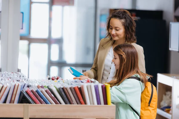 Woman smiling near daughter choosing new copybook in stationery shop — Stock Photo