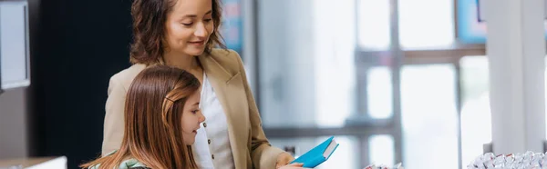 Smiling woman showing new copybook to daughter in stationery store, banner — стоковое фото