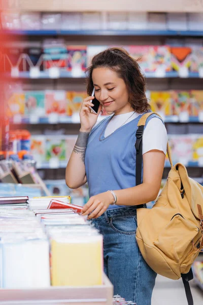 Усміхнена жінка розмовляє на мобільному телефоні при виборі з різноманітних книг у магазині — стокове фото