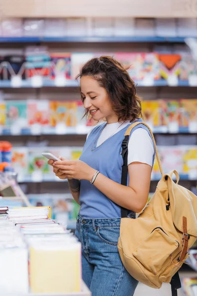 Smiling student with backpack messaging on mobile phone in blurred stationery store — Fotografia de Stock